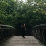 A woman with curly hair, wearing a black sleeveless outfit, walks alone across a wooden footbridge surrounded by dense green trees in Collingwood, Ontario. The overcast sky above adds a quiet, introspective atmosphere to the scene as she heads toward the forest.