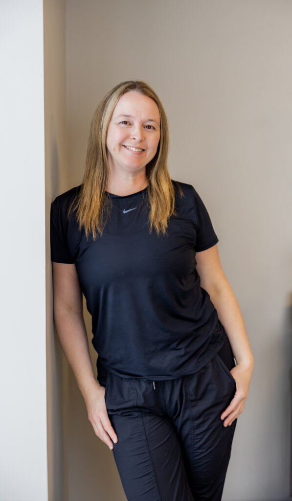 April Markou standing against a light wall with one hand in her pocket. She wears a black Nike top and black pants, smiling in a relaxed and confident posture.