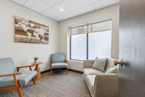 A cozy consultation or waiting room with a small sofa and two light blue armchairs around a wooden side table. A wood and resin world map hangs above the chairs, with frosted windows letting in soft light.