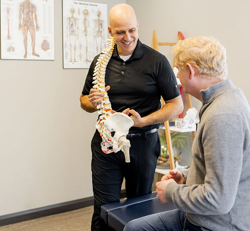 Dr. Peter Wilson holding a spine model while explaining it to a client seated on a bench in a clinical room. Anatomy posters and a window bring light into the space.