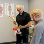 Dr. Peter Wilson holding a detailed spine model and explaining its anatomy to a blonde male patient. Anatomical charts line the wall behind him.