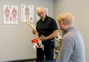 Dr. Peter Wilson holding a detailed spine model and explaining its anatomy to a blonde male patient. Anatomical charts line the wall behind him.