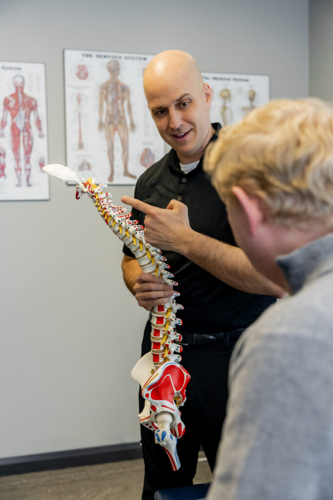 Close-up of Dr. Peter Wilson gesturing toward a spine model while discussing vertebrae and nerve pathways with a client. Educational posters are visible in the background.
