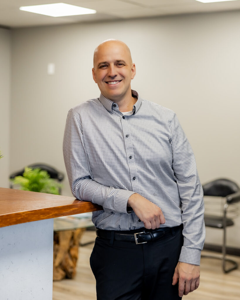 Dr. Peter Wilson leaning casually against the front counter, smiling at the camera. He’s dressed in a checkered button-up shirt and black trousers.
