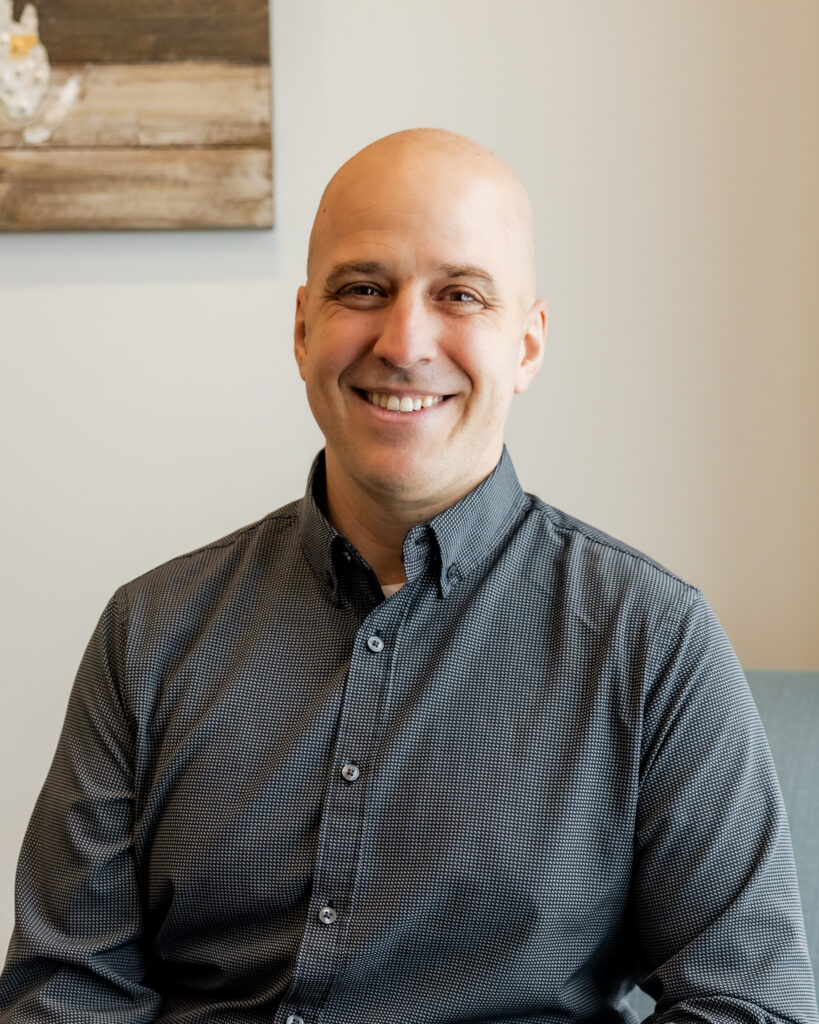 Dr. Peter Wilson seated on a light blue armchair, smiling and relaxed. He wears a grey checkered shirt with a rustic wood wall art piece behind him.