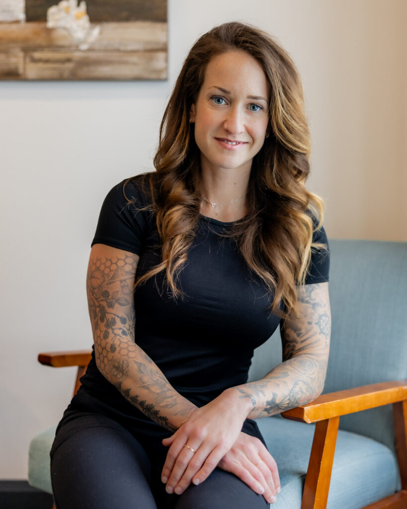 Sophie Szrajer seated on a light blue armchair, smiling with her hands resting in her lap. A wood and resin wall art piece is visible behind her.