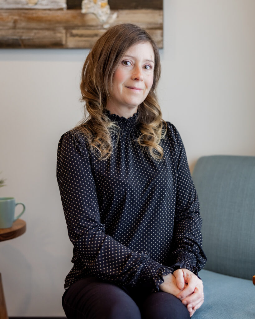 Tanya Lawrence seated in a light blue armchair, smiling with her hands folded in her lap. She wears a black blouse with white polka dots, and a wood and resin wall art piece is visible behind her.