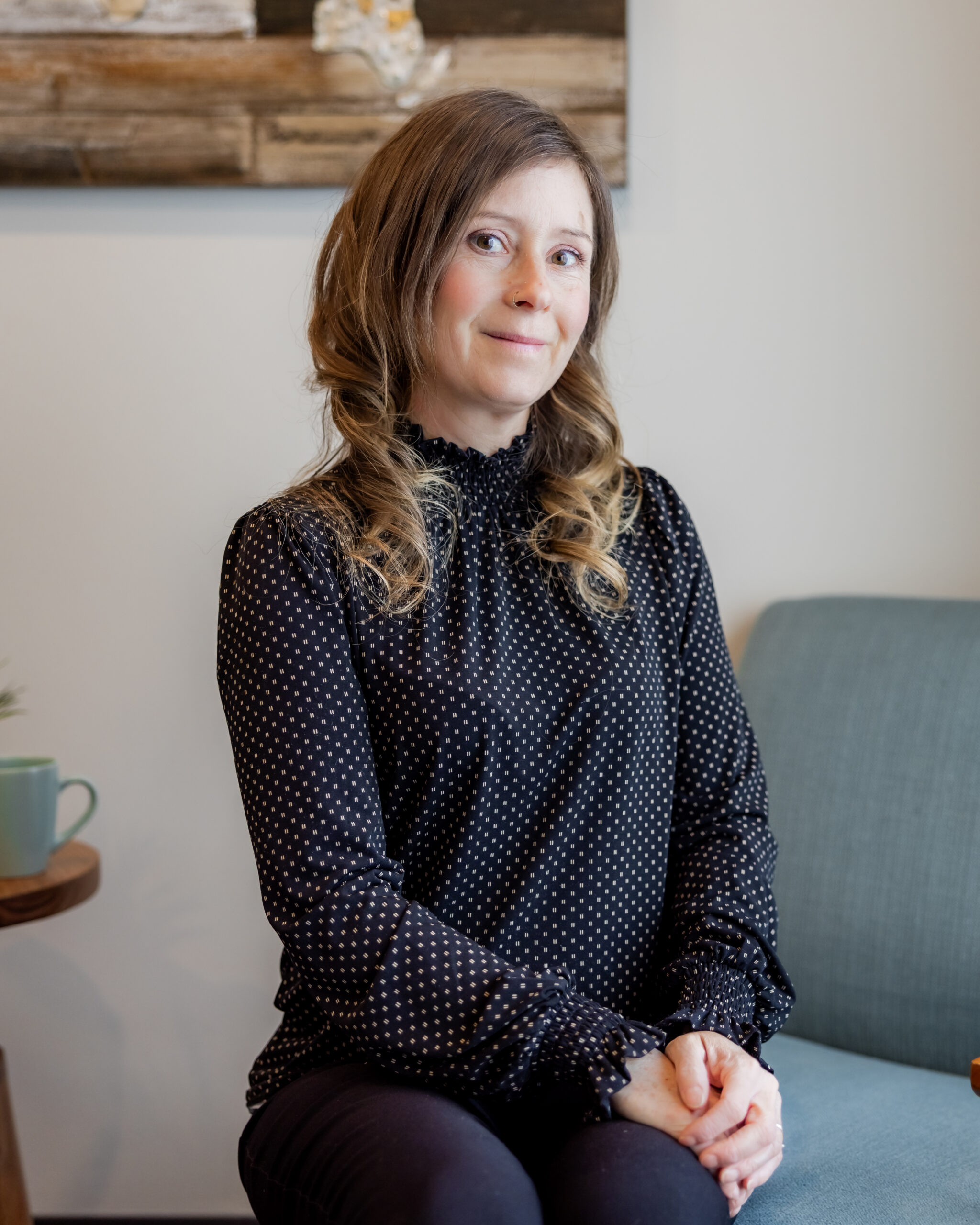 Tanya Lawrence seated in a light blue armchair, smiling with her hands folded in her lap. She wears a black blouse with white polka dots, and a wood and resin wall art piece is visible behind her.