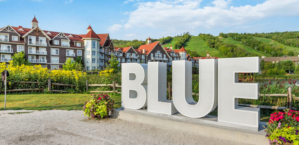 Large white "BLUE" monument sign surrounded by colorful flowers and landscaping, with elegant chalet-style resort buildings featuring red roofs and timber accents in the background. Green ski hills and forest visible behind the resort under a bright blue sky.