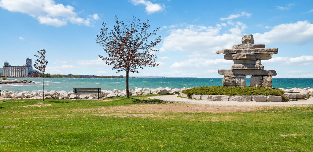 Waterfront park featuring a traditional inukshuk stone monument on the right, with a bench and young tree on the left. Rocky shoreline borders turquoise blue water, with a grain elevator visible in the distance under a bright blue sky with scattered clouds.