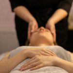 Person lying on a massage table with their hands resting over a towel-covered torso, while a practitioner performs a gentle head or cranial massage. The setting suggests a calm, professional wellness or spa environment. Photo by Anna Keibalo on Unsplash