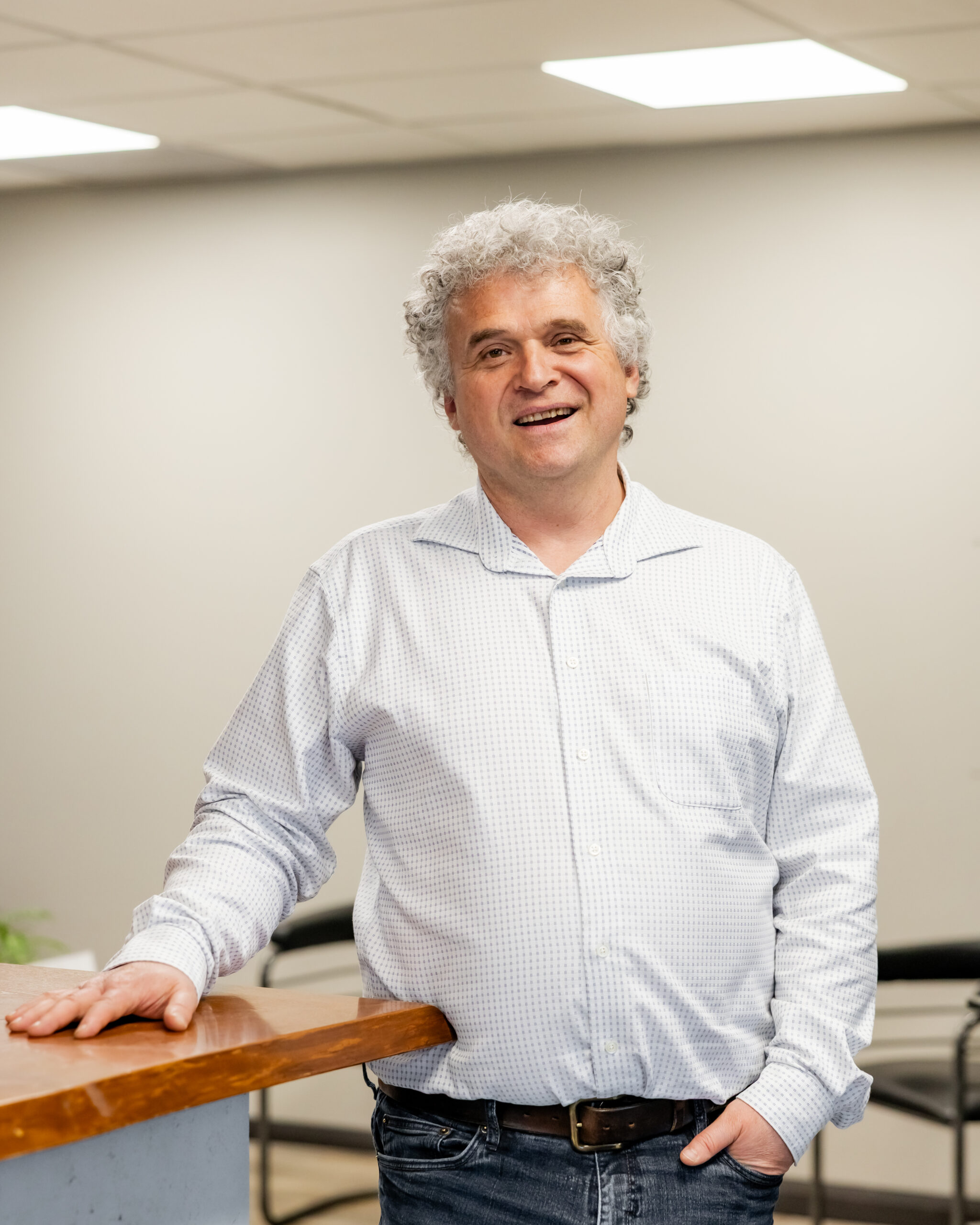 Kaleb Einstein Montgomery standing beside a wood and glass reception counter, smiling with one hand resting on the counter. He wears a light blue patterned shirt and jeans.