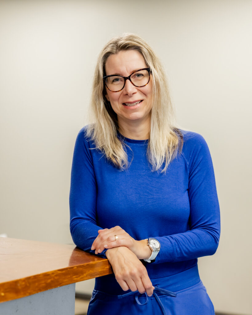Mirka Grell-Willins leaning casually against a wood reception counter, smiling. She wears a bright blue long-sleeved top and matching pants, glasses, and a watch.