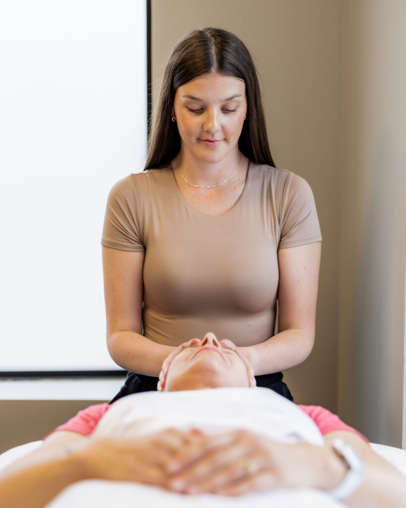 Rachel Cubitt performing craniosacral therapy, gently holding a patient's head with both hands while the patient lies on their back.