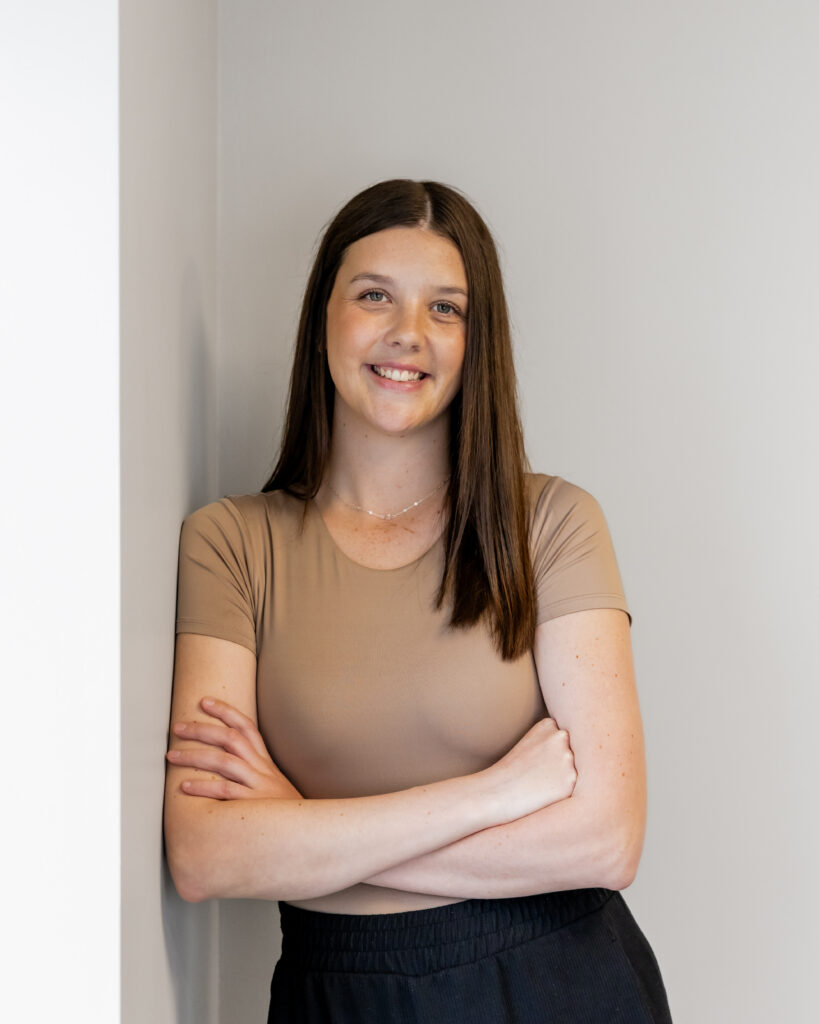 Rachel Cubitt standing against a light-colored wall, smiling with arms crossed. She wears a short-sleeved taupe top and black pants.